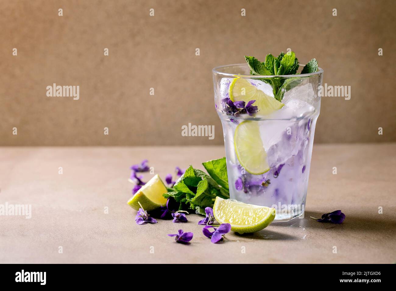 Verre transparent de soda à la limonade florale avec menthe, glace à la fleur de violettes et rondelles de lime sur une table brune. Rafraîchissement d'été froid Banque D'Images