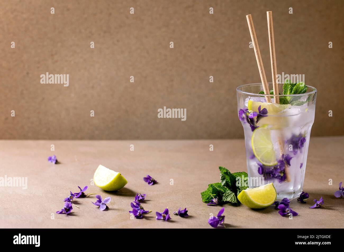 Verre transparent de soda à la limonade avec menthe, glace à la fleur de violettes et tranches de lime, pailles en papier, debout sur une table brune. Boisson d'été froide Banque D'Images