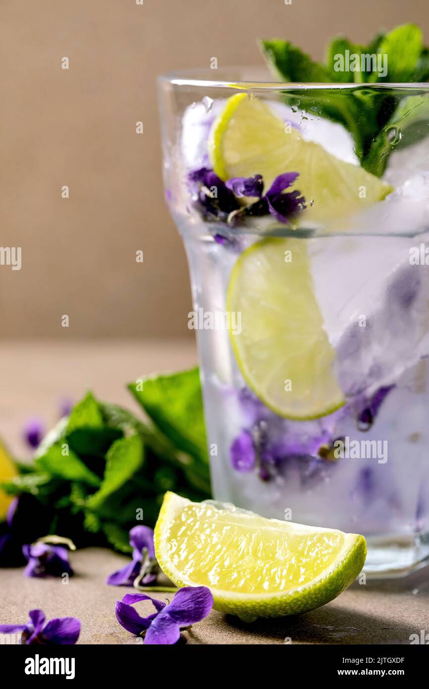 Verre transparent de soda à la limonade florale avec menthe, glace à la fleur de violettes et rondelles de lime sur une table brune. Rafraîchissement d'été froid Banque D'Images