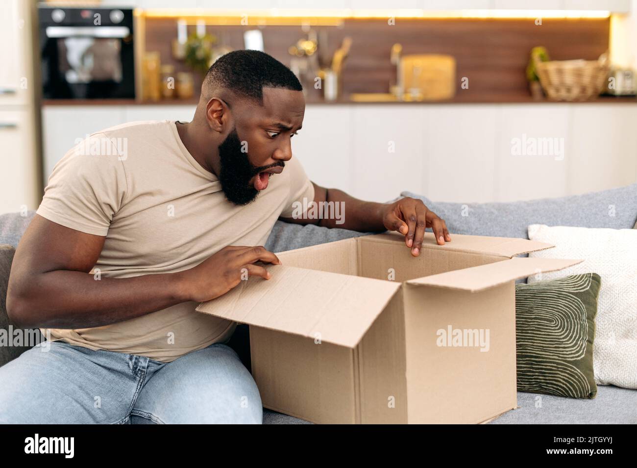 Un gars afro-américain confus choqué ouvrant une grande boîte de carton, déballant la commande d'un magasin Internet à la maison, se sent frustré par le service de livraison longue et les marchandises qui ont reçu, éprouvant Banque D'Images