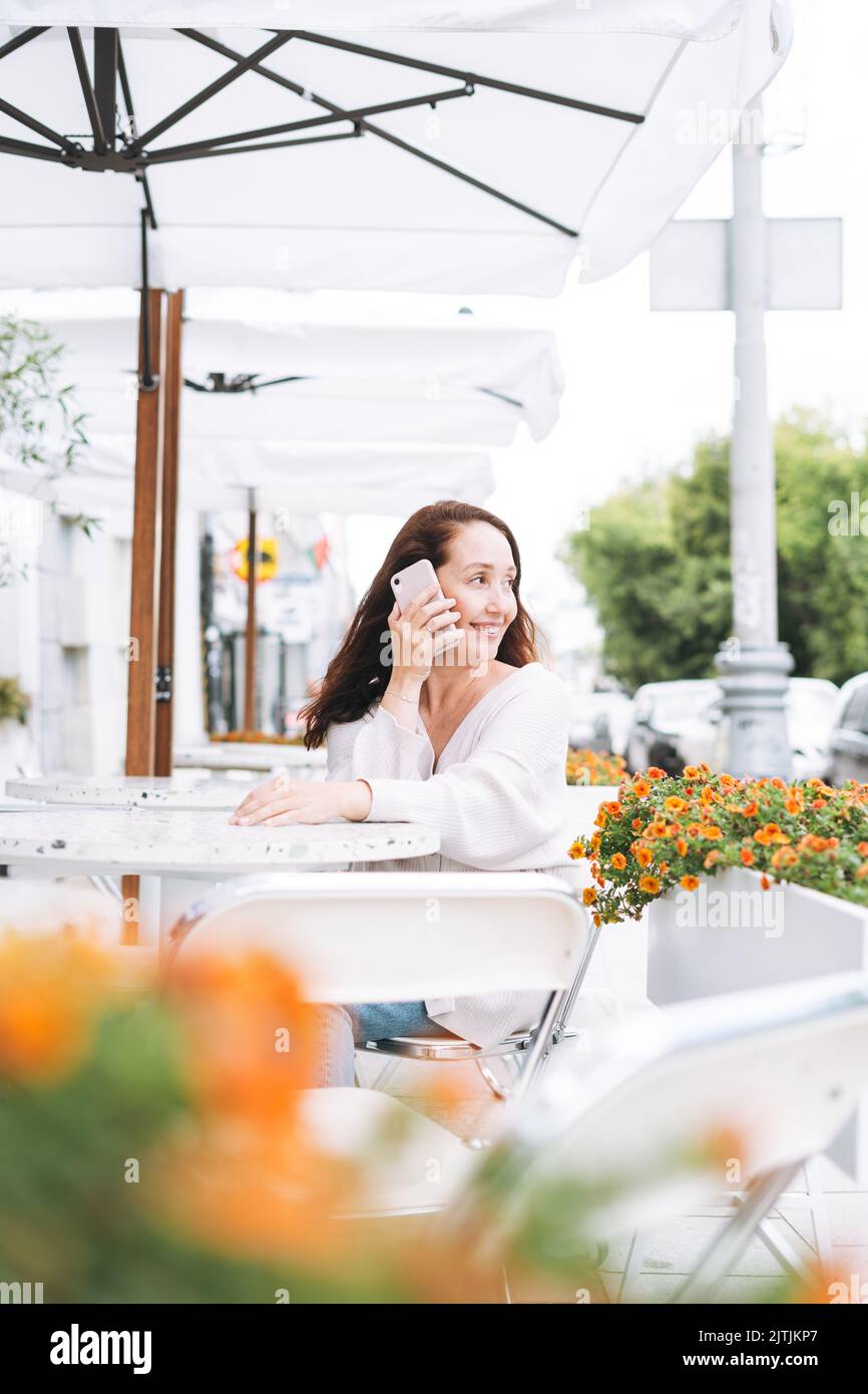 Femme brune souriante adulte quarante ans utilisant un téléphone portable dans un café de la rue de la ville Banque D'Images