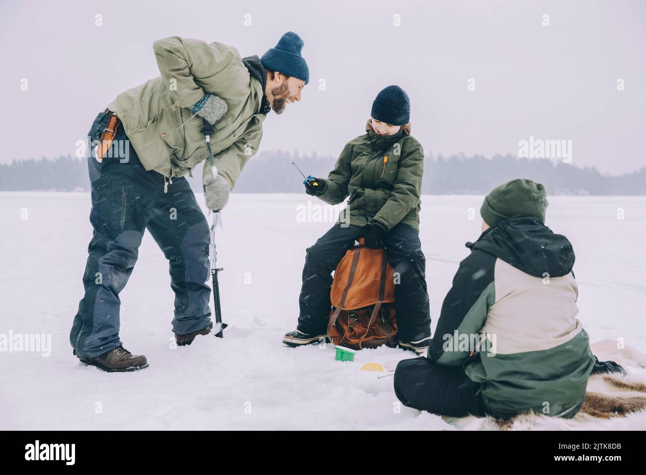 Homme joyeux utilisant la tarière de glace tout en parlant avec sac à dos au lac gelé Banque D'Images