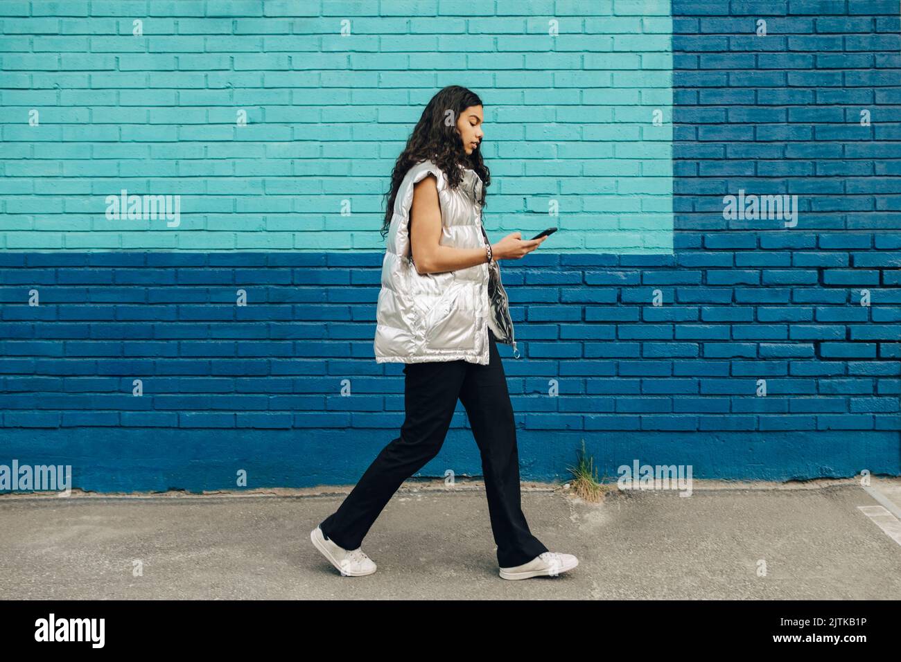 Une jeune femme utilisant un téléphone portable marchant près d'un mur de briques bleues Banque D'Images