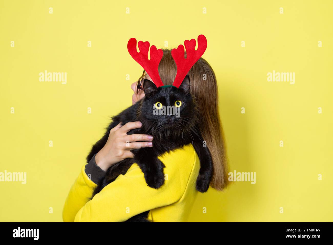 Fille dans une robe jaune tient son bien-aimé doux chat noir drôle avec les yeux jaunes dans le bandeau de Noël renne Antlers sur un fond jaune Banque D'Images