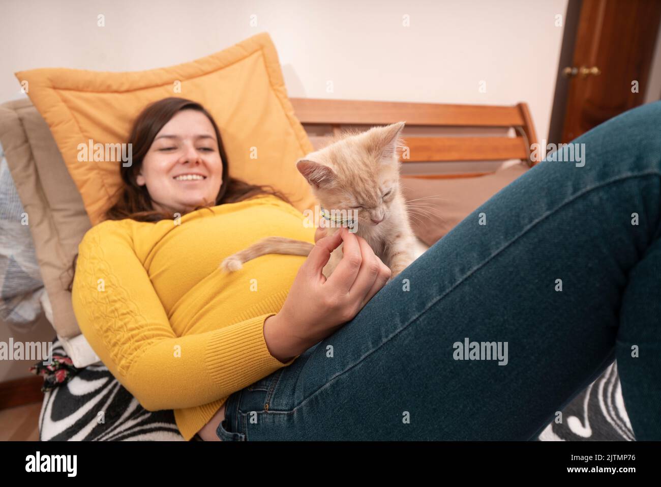 Belle jeune femme hispanique souriante vêtue d'une veste jaune, couchée sur son lit en jouant avec son petit chaton. Chat concentré Banque D'Images