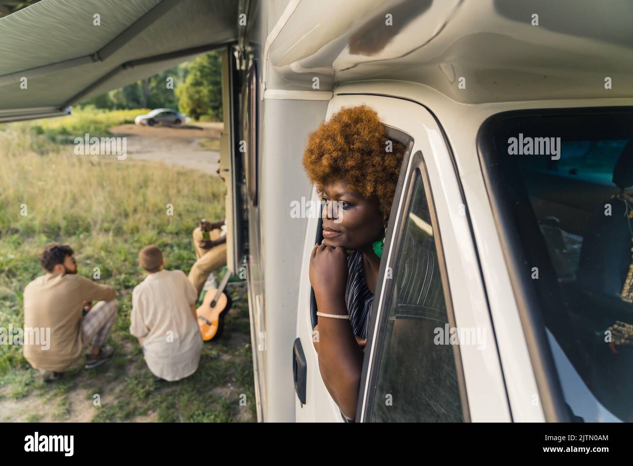 Jeune femme africaine à la peau sombre et attentionnés avec des cheveux kinésitants, penchée par la fenêtre d'une camionnette et ses amis assis sur l'herbe à côté d'elle à la fin de leur voyage. Photo de haute qualité Banque D'Images