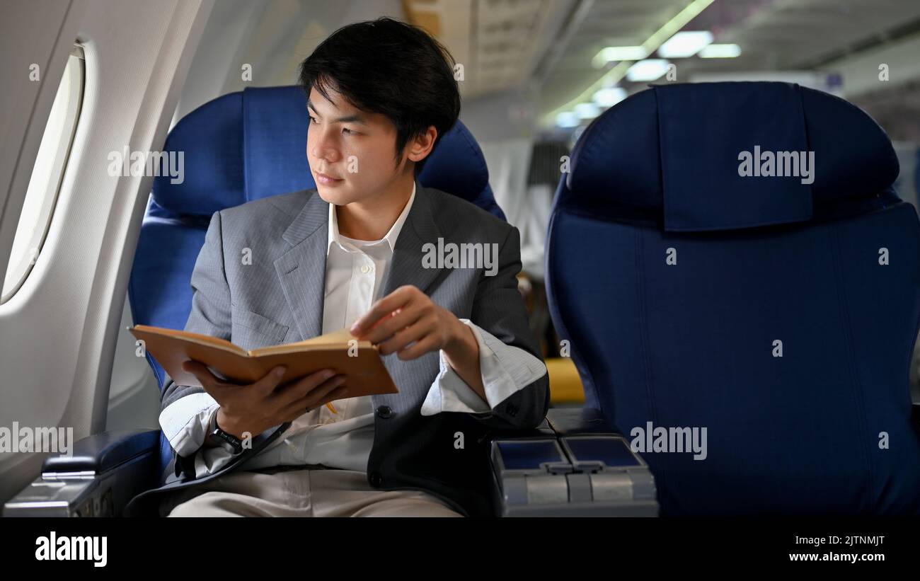 Un jeune homme d'affaires asiatique ou un directeur général homme à succès tient un livre tout en regardant la fenêtre, lisant un livre pendant le vol pour son bus Banque D'Images