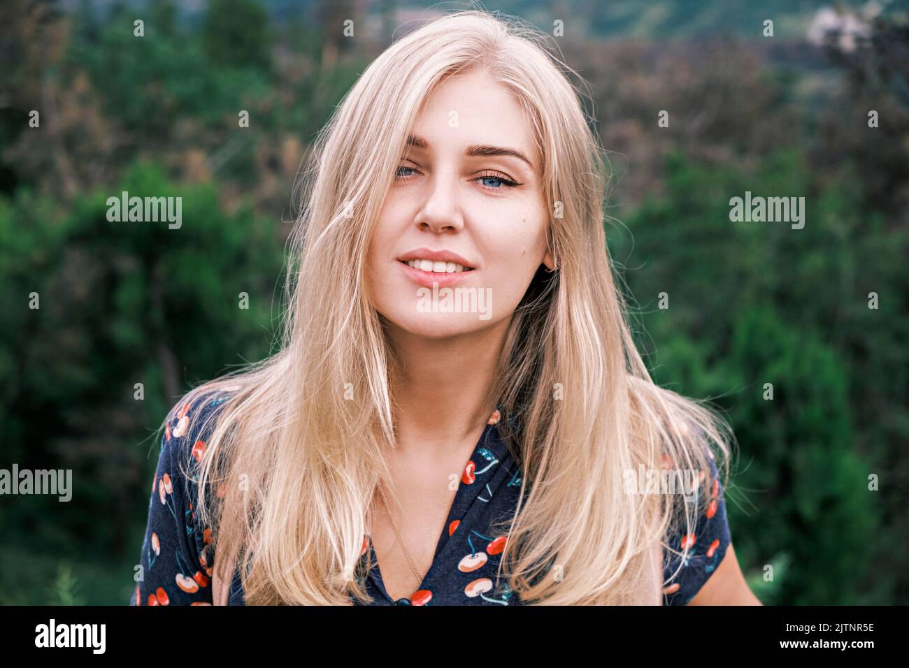 Portrait d'une jeune femme blonde aux yeux bleus avec de longs cheveux regardant dans l'appareil photo et souriant devant un arrière-plan flou en extérieur Banque D'Images