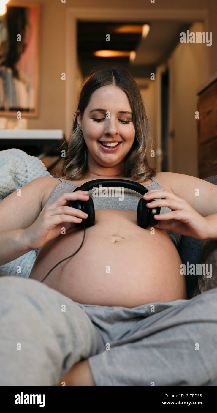 Gros plan sur une maman enceinte assise sur son canapé en souriant en écoutant de la musique avec un casque Banque D'Images