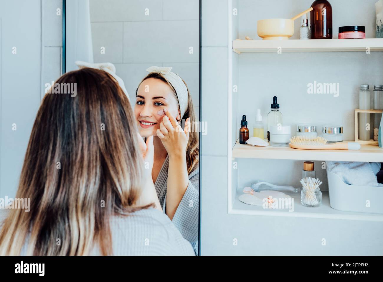 Jeune femme en peignoir regardant dans le miroir et appliquant le masque en argile cosmétique naturel du visage sur son visage dans la salle de bains. Procédures cosmétiques pour voiture de peau Banque D'Images Jeune femme en peignoir regardant dans le miroir et appliquant le masque en argile cosmétique naturel du visage sur son visage dans la salle de bains. Procédures cosmétiques pour voiture de peau Banque D'Images