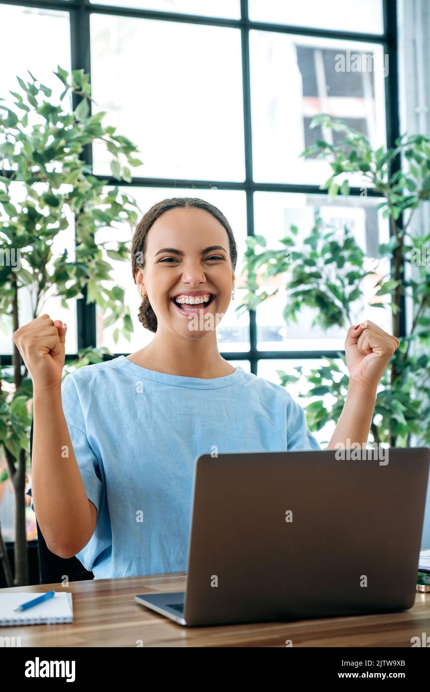 Photo verticale d'une jeune femme d'affaires heureuse et heureuse, assis à un bureau avec un ordinateur portable au bureau, se réjouir du succès, gros profit, célébrer l'affaire, faire des gestes avec les poings, regarde l'appareil photo, souriant Banque D'Images