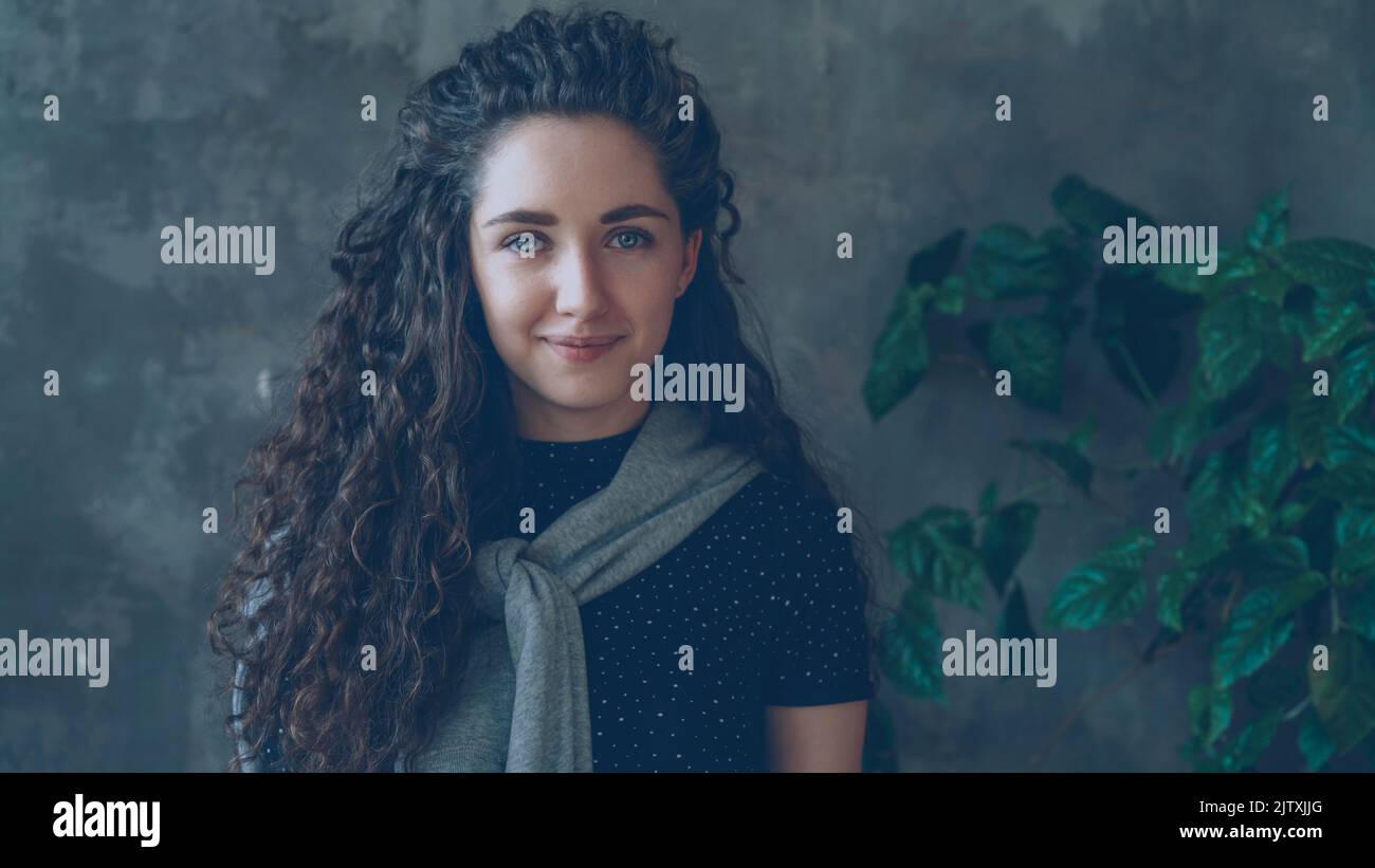 Portrait en gros plan de jeune fille attrayante regardant dans l'appareil photo et souriant tout en se tenant contre le mur gris et la grande plante verte à l'intérieur Banque D'Images
