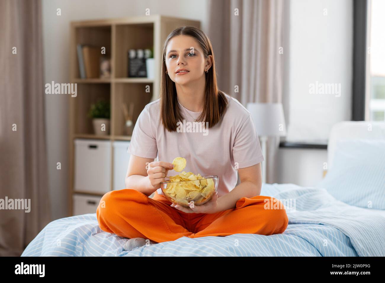 bonne fille mangeant des chips assis sur le lit à la maison Banque D'Images