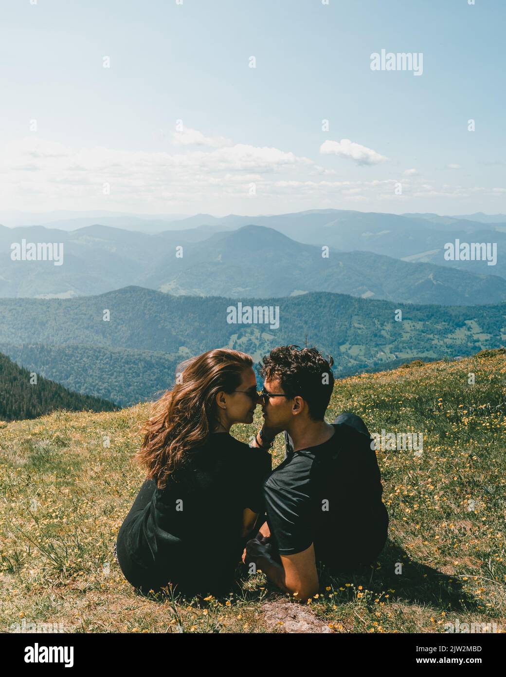 Vue arrière d'un jeune couple romantique en t-shirts noirs et lunettes de soleil embrassant tout en étant assis sur un pré herbacé avec vue pittoresque sur la montagne pendant le voyage Banque D'Images Vue arrière d'un jeune couple romantique en t-shirts noirs et lunettes de soleil embrassant tout en étant assis sur un pré herbacé avec vue pittoresque sur la montagne pendant le voyage Banque D'Images