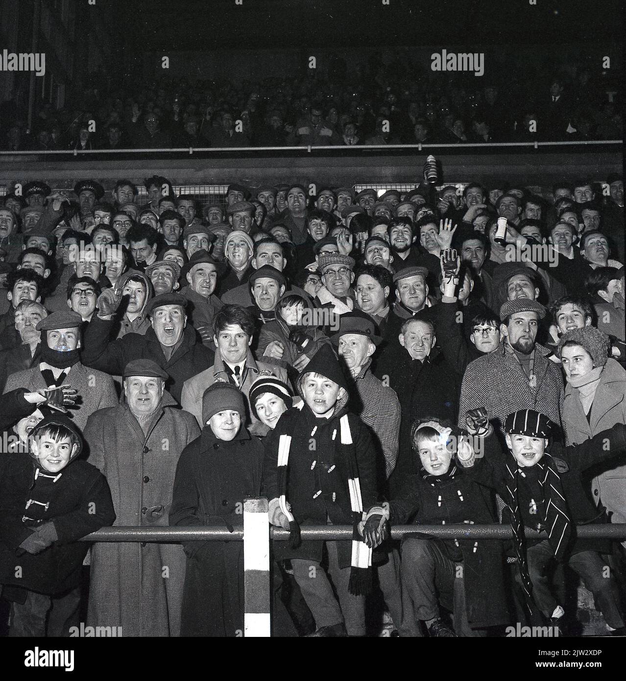 1960s, des fans de football historiques se tenant ensemble sur une terrasse traditionnelle pour regarder un match, lors d'un match en soirée... manteaux, casquettes plates pour hommes, manteaux de sport et chapeaux de galet pour les jeunes, l'époque avant les répliques de chemises! Banque D'Images