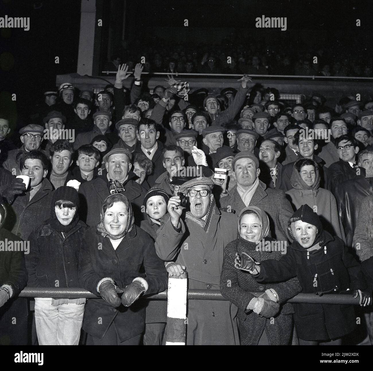1960s, des fans de football historiques se tenant ensemble sur une terrasse traditionnelle pour regarder un match, lors d'un match en soirée... manteaux, casquettes plates pour hommes, manteaux de sport et chapeaux de galet pour les jeunes, l'époque avant les répliques de chemises! Banque D'Images