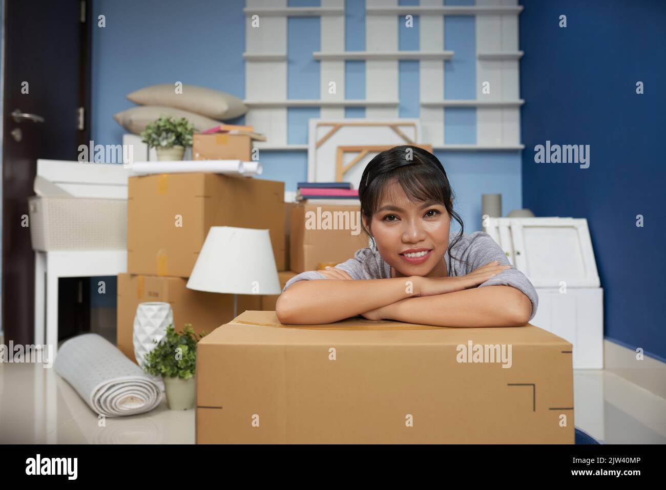 Portrait d'une jeune femme asiatique penchée sur une boîte en carton et souriant à l'appareil photo tout en se défaisant dans un nouvel appartement Banque D'Images