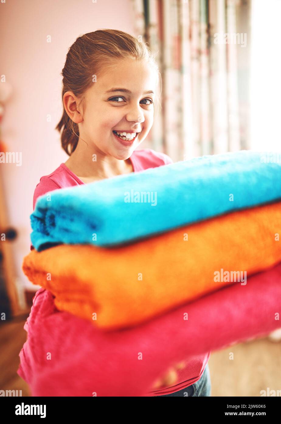 Tout propre et doux. Portrait d'une petite fille gaie portant un tas de serviettes fraîchement lavées tout en regardant l'appareil photo à la maison. Banque D'Images