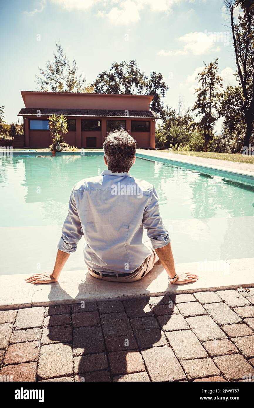 Un jeune homme vu de derrière est assis près de la piscine. Homme d'affaires prend un moment de détente au bord de la piscine. Banque D'Images