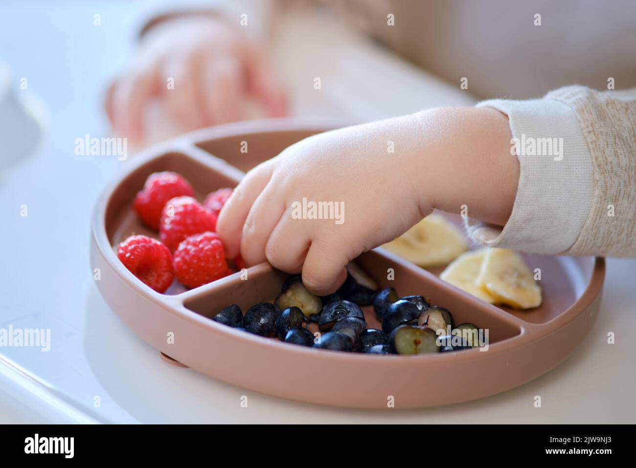 Bébé mange des fruits et des baies avec sa main, table en gros plan. Les mains des enfants prennent les aliments d'une assiette beige. Enfant âgé d'un an et deux mois Banque D'Images