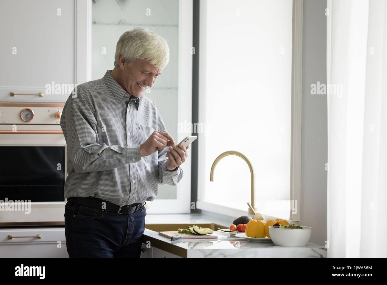 Joyeux homme retraité plus âgé utilisant un téléphone portable sur le comptoir de la cuisine Banque D'Images