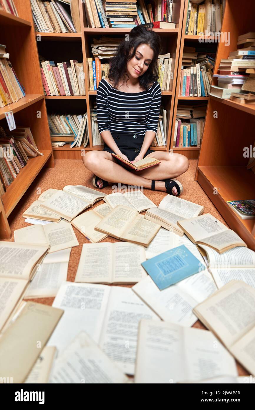 Belle jeune femme en haut rayé et mini jupe noire dans une bibliothèque pleine de livres, posant comme un bibliothécaire ou étudiant Banque D'Images