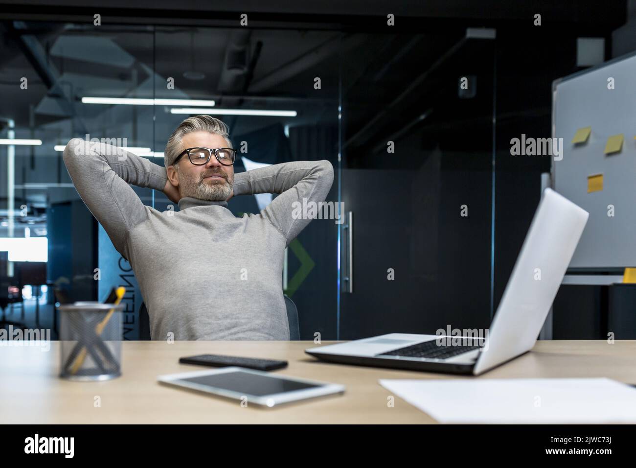 Homme d'affaires senior aux cheveux gris reposant au bureau sur une chaise avec les mains derrière la tête, chef d'entreprise travaillant avec un ordinateur portable, investisseur mature a terminé le projet, rêver. Banque D'Images