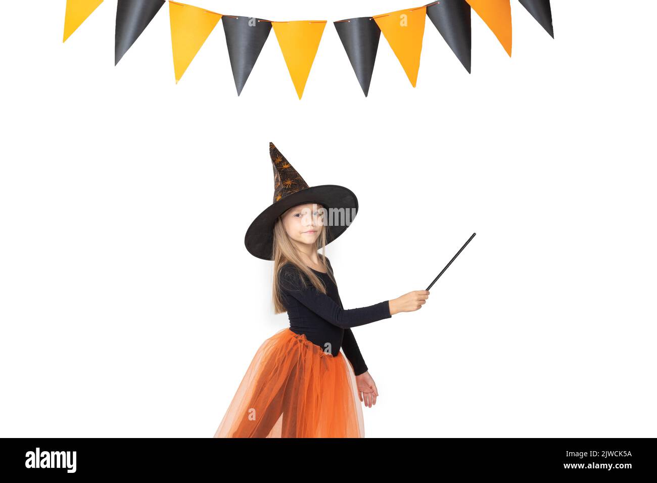 Halloween pour enfants. Une jolie petite fille dans un costume de sorcière, portant un chapeau, tenant une baguette magique montre un espace vide sur un fond blanc avec un garlan Banque D'Images