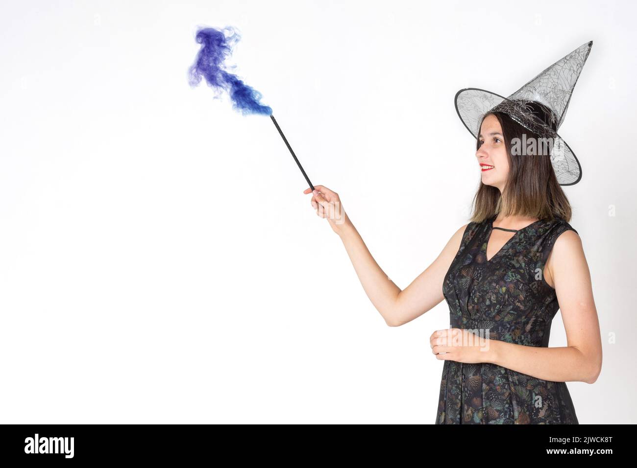 Une belle jeune fille dans un costume de sorcière, portant un chapeau, avec une baguette magique avec la fumée tourbillonnante colorée. La sorcière est attrayante. Joyeux Halloween. Banque D'Images