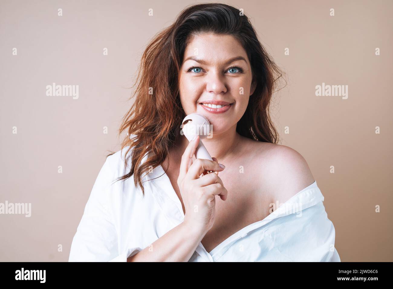 Auto-affectueuse femme plus taille en chemise blanche avec masseur facial dans les mains sur fond beige, amour du corps Banque D'Images