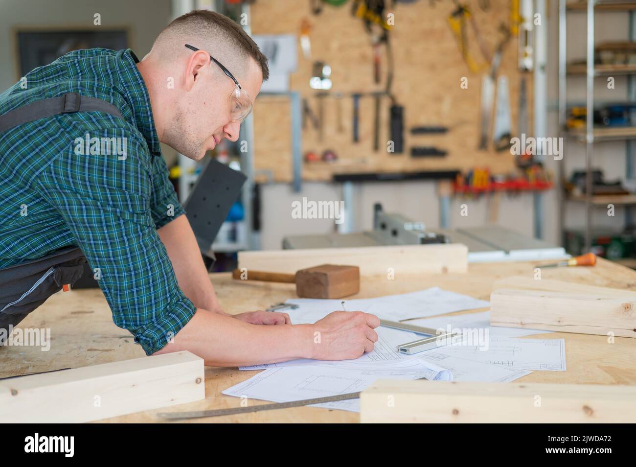 Un portrait d'un menuisier dans des lunettes et des combinaisons de travail dessine un plan pour un atelier. Banque D'Images