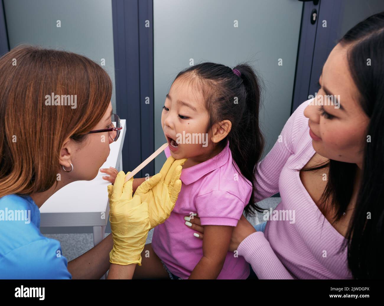 Médecin utilisant une spatule d'inspection pour examiner la gorge de la petite fille coréenne. Mignonne fille avec sa mère au rendez-vous du pédiatre Banque D'Images