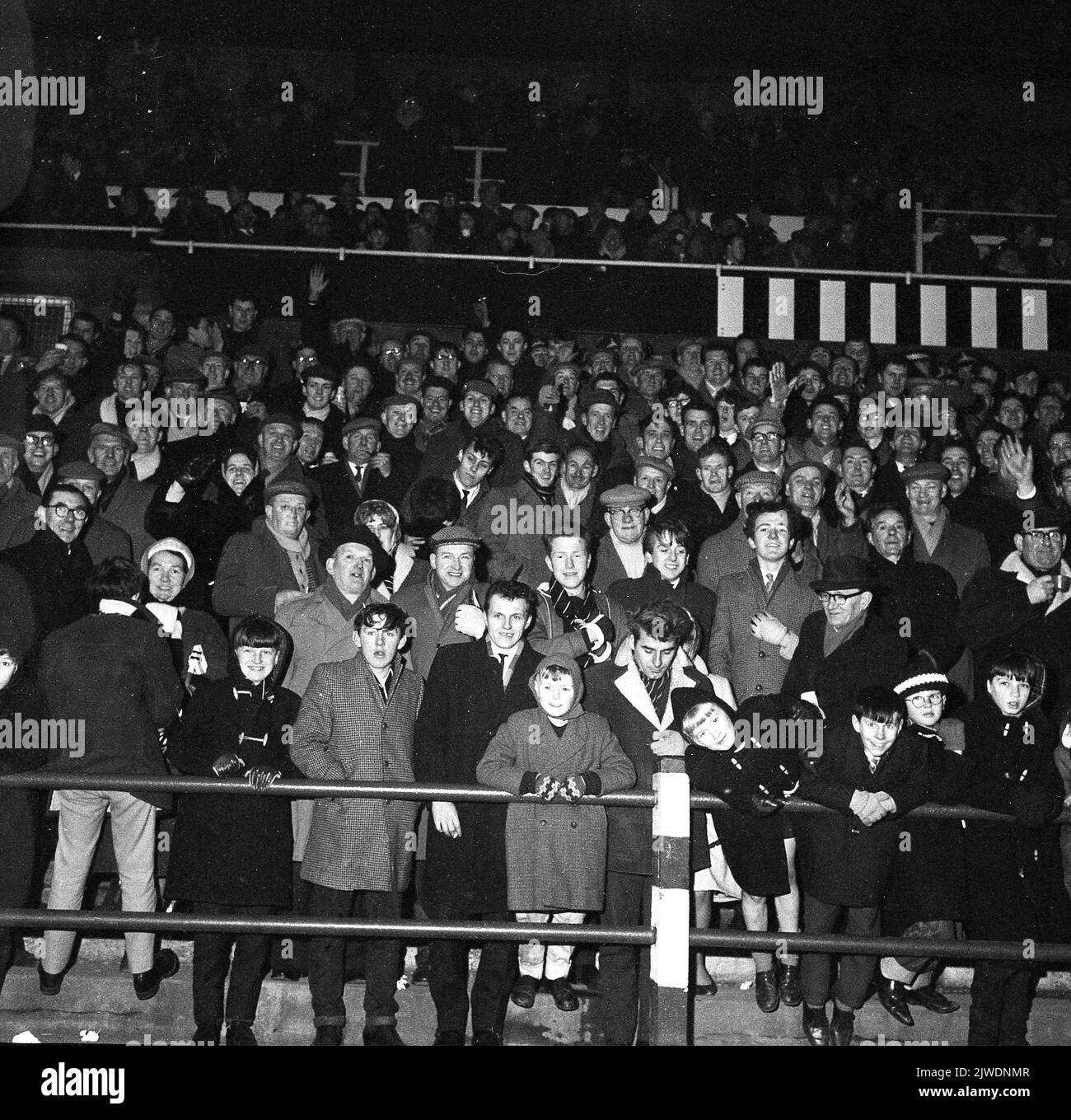 1960s, des fans de football historiques se tenant ensemble sur les terrasses pour un match de soirée... manteaux, casquettes plates pour les hommes, manteaux de sport et chapeaux de galet pour les jeunes, pas une réplique de chemise en vue! Banque D'Images