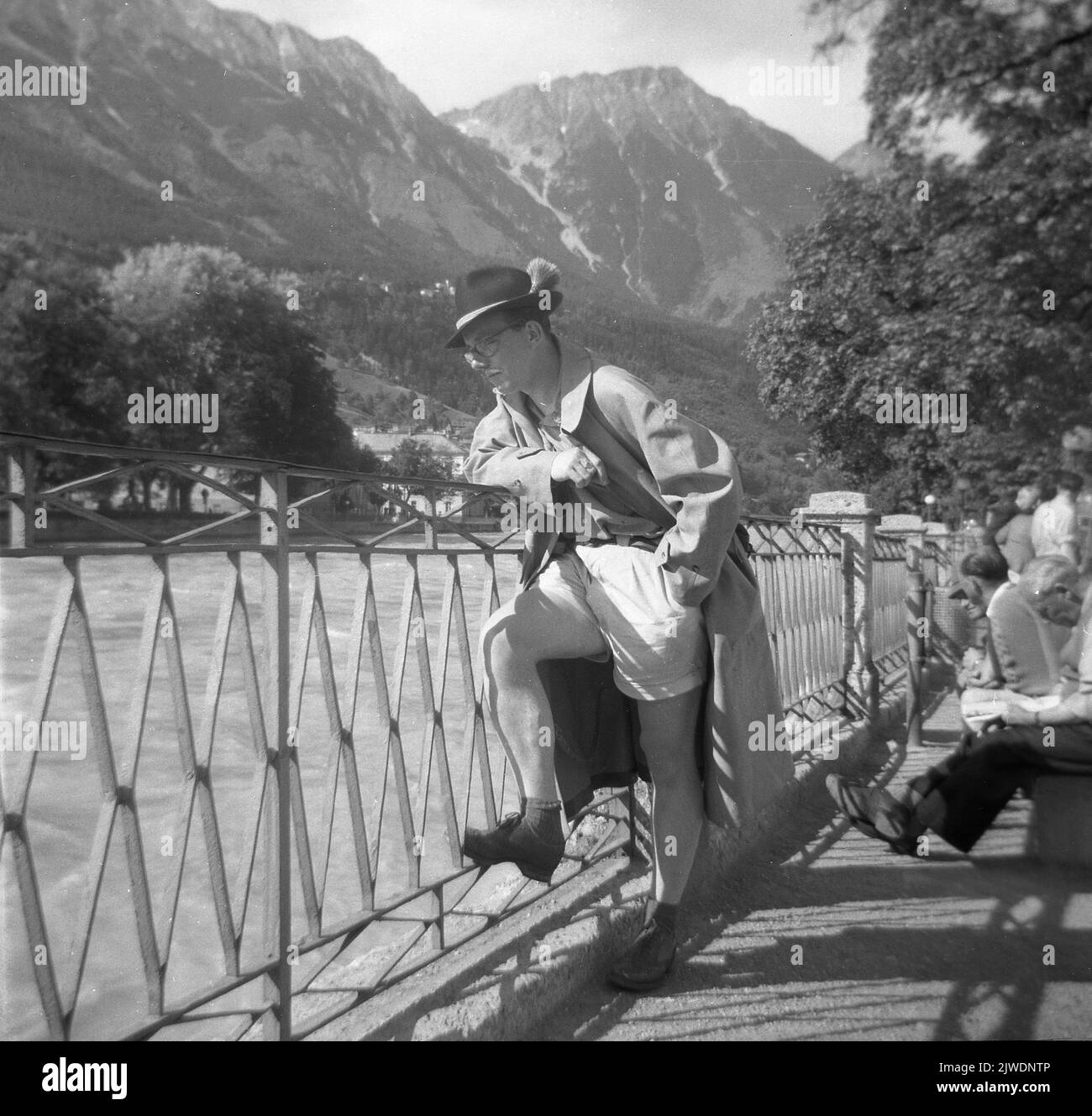 1958, historique, un touriste anglais en short, pardessus, et chapeau alpin traditionnel avec plumes, debout près d'une barrière qui regarde sur une rivière dans la région de Tryol en Autriche. Les Alpes en arrière-plan. Banque D'Images