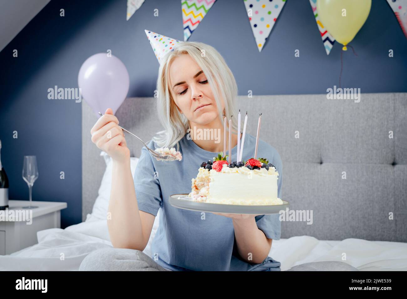 Triste, bouleversée femme en pajama et tête de fête pensant manger ou de ne pas manger gâteau d'anniversaire assis sur le lit dans la chambre décorée. Fête son anniversaire seul c Banque D'Images Triste, bouleversée femme en pajama et tête de fête pensant manger ou de ne pas manger gâteau d'anniversaire assis sur le lit dans la chambre décorée. Fête son anniversaire seul c Banque D'Images