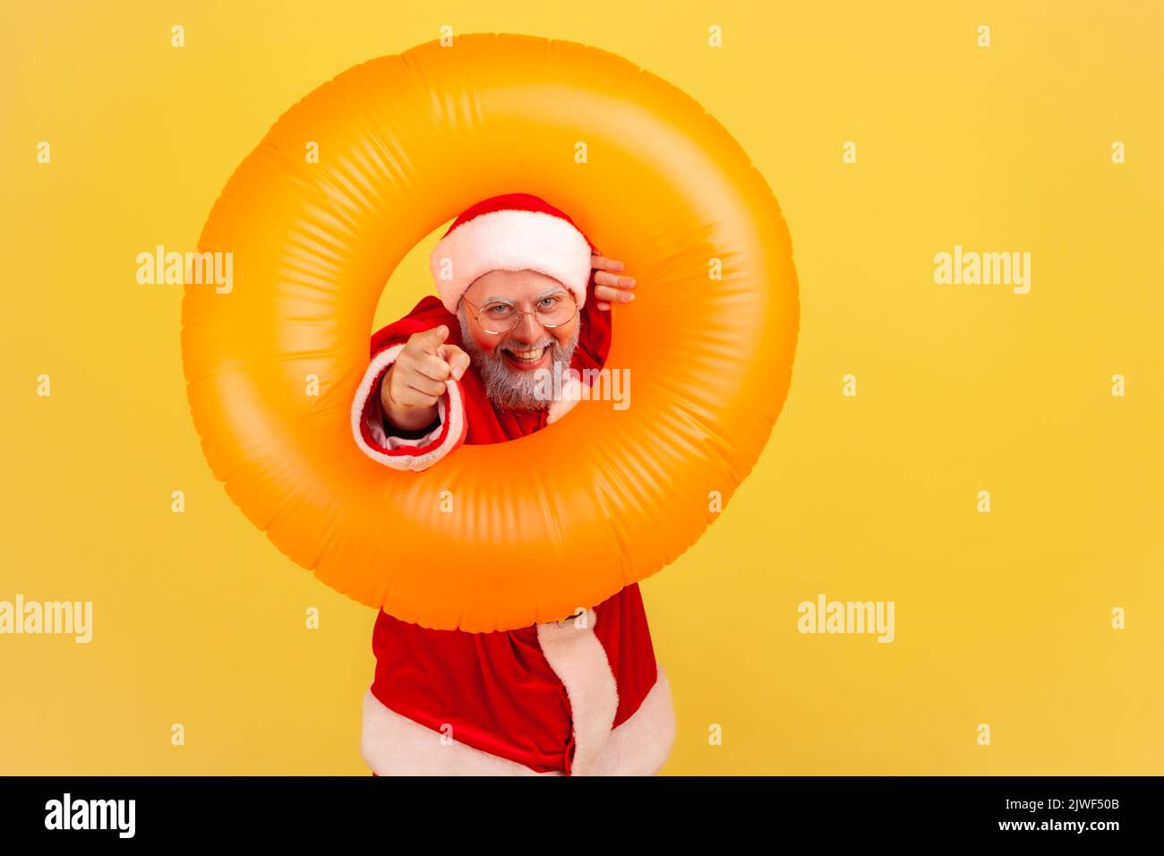 Joyeux homme âgé avec une barbe grise en costume du père noël debout avec anneau en caoutchouc, pointant vers l'appareil photo avec un sourire crasseux, ayant une expression positive. Studio d'intérieur isolé sur fond jaune. Banque D'Images