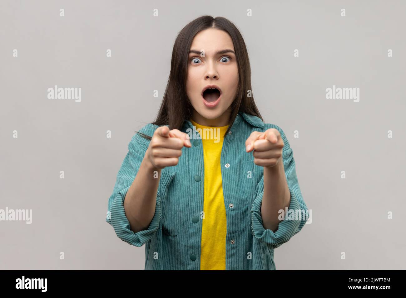 Femme surprise avec des cheveux foncés pointant vers l'appareil photo avec une expression surprise choquée, faisant le choix, portant une veste de style décontracté. Prise de vue en studio isolée sur fond gris. Banque D'Images