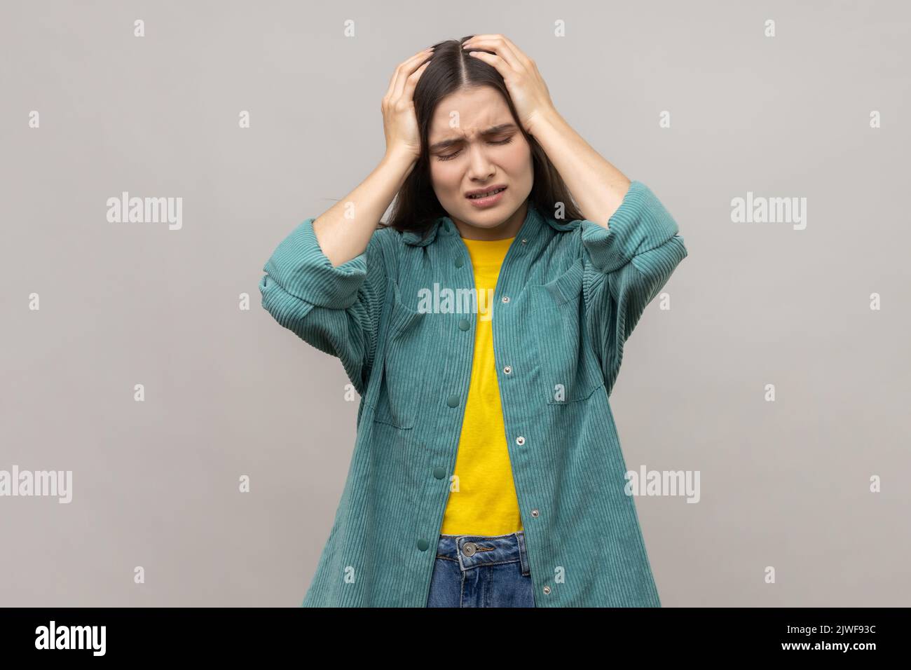 Portrait d'une femme malade malsaine aux cheveux foncés tenant les mains sur la tête, se sentant mal de tête terrible, risque de migraine, portant une veste de style décontracté. Prise de vue en studio isolée sur fond gris. Banque D'Images