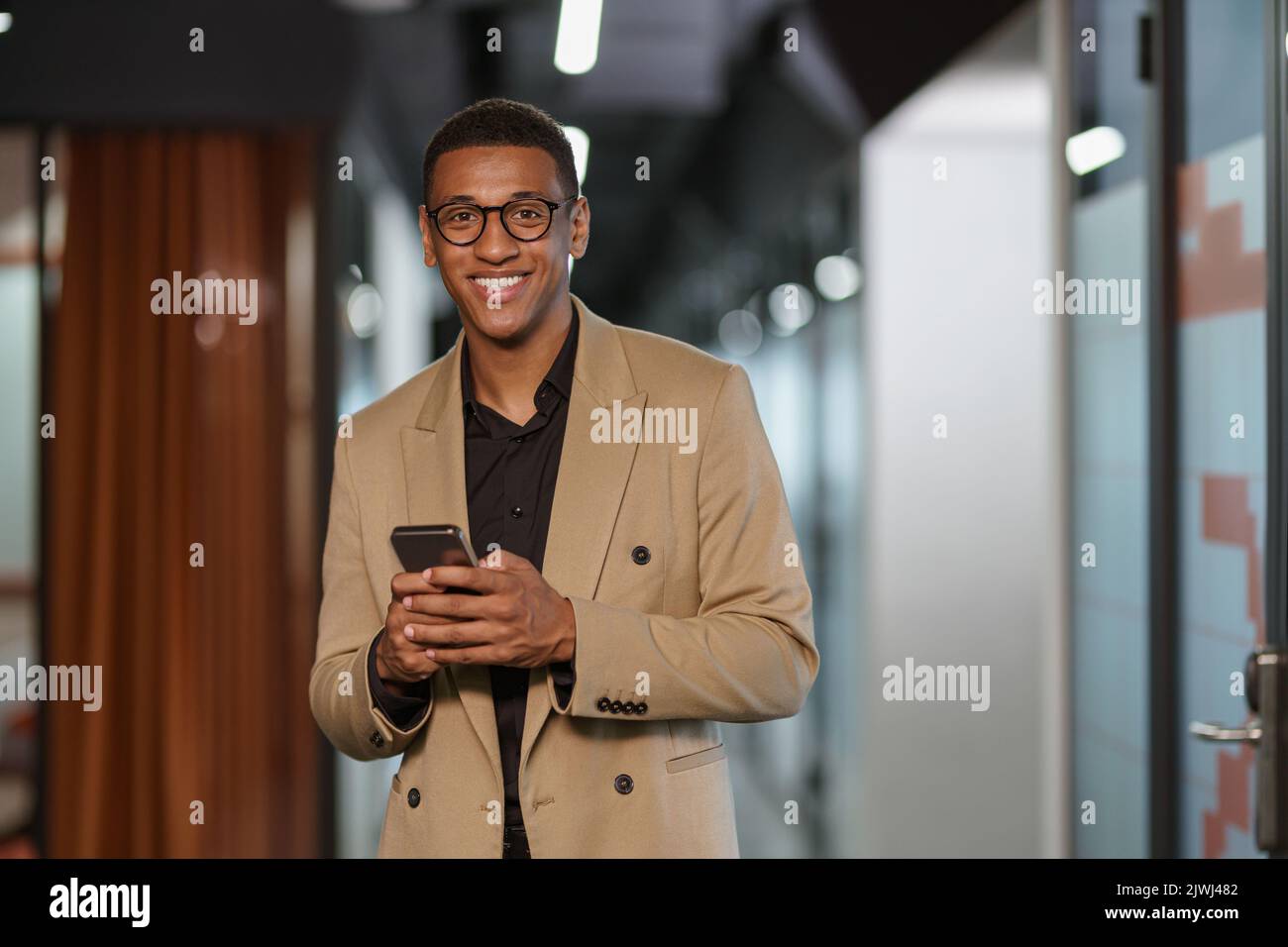 Beau homme d'affaires africain souriant utilisant le téléphone tout en se tenant dans un collègue moderne Banque D'Images