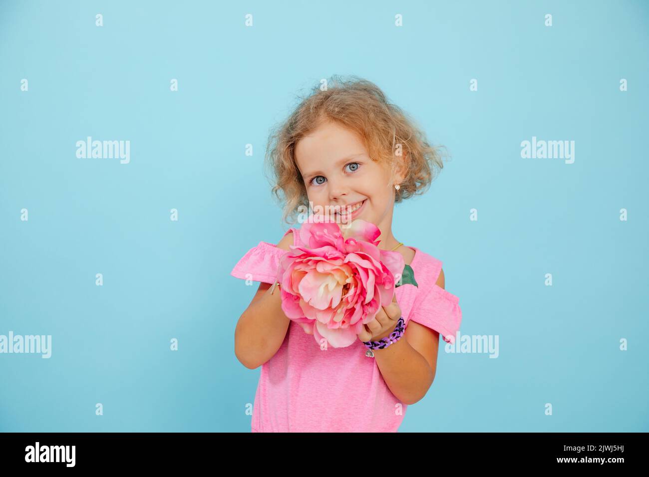 Charmante petite fille montre beau métier floral sur fond bleu vide, espace libre de copie. Enfant à yeux bleus de l'âge de la maternelle tenir le papier fleur rose Banque D'Images