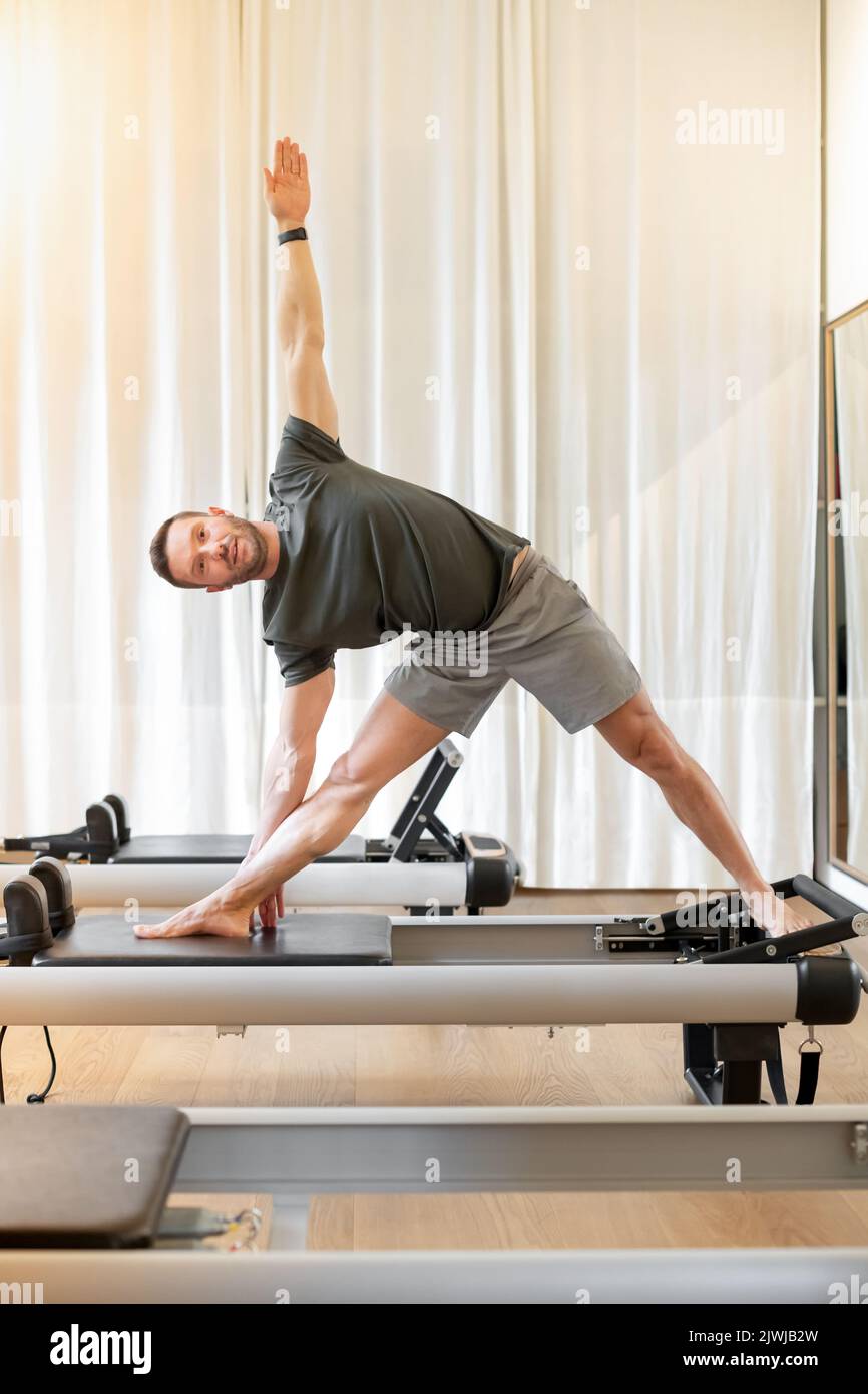 Corps complet de sportive pieds nus mâle en activewear pratiquant triangle poser sur le lit reformeur pendant l'entraînement pilates dans la salle de gym moderne Banque D'Images