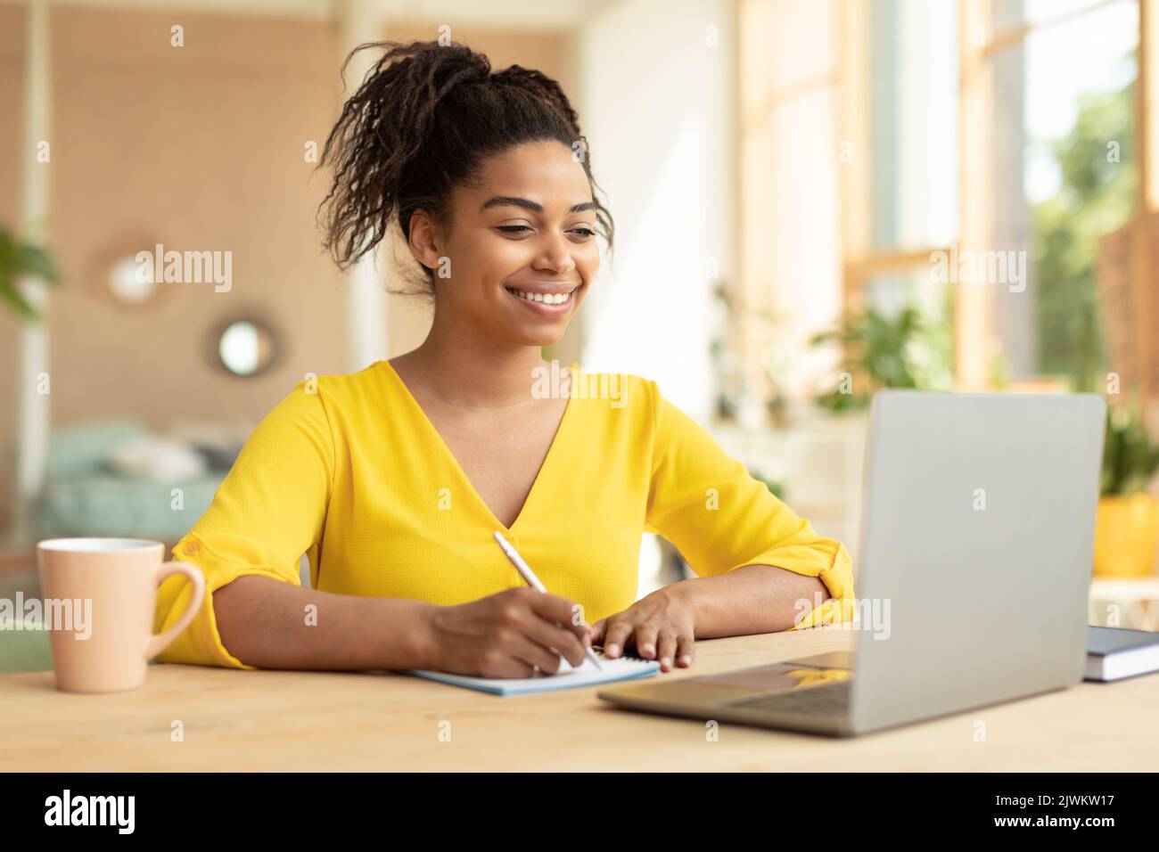 Femme afro-américaine souriante travaillant sur un ordinateur portable prenant des notes dans un ordinateur portable, assise au bureau à la maison, espace de copie Banque D'Images