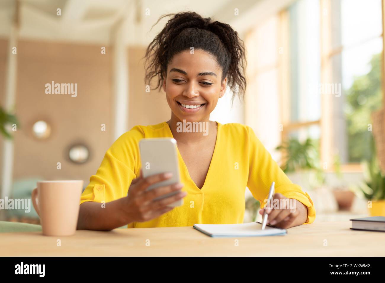 Apprentissage à distance. Bonne femme noire utilisant un smartphone et écrivant dans son ordinateur portable, regardant des conférences en ligne à la maison Banque D'Images