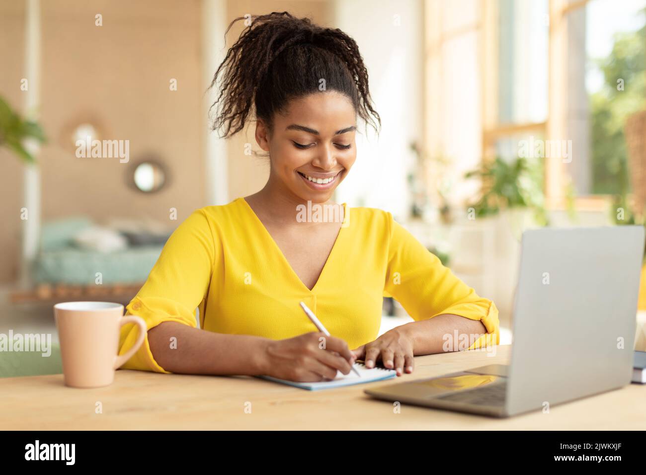 Concept de prise de notes. Jeune femme noire heureuse utilisant un ordinateur portable et écrivant dans son cahier d'exercices, assis au bureau à la maison Banque D'Images