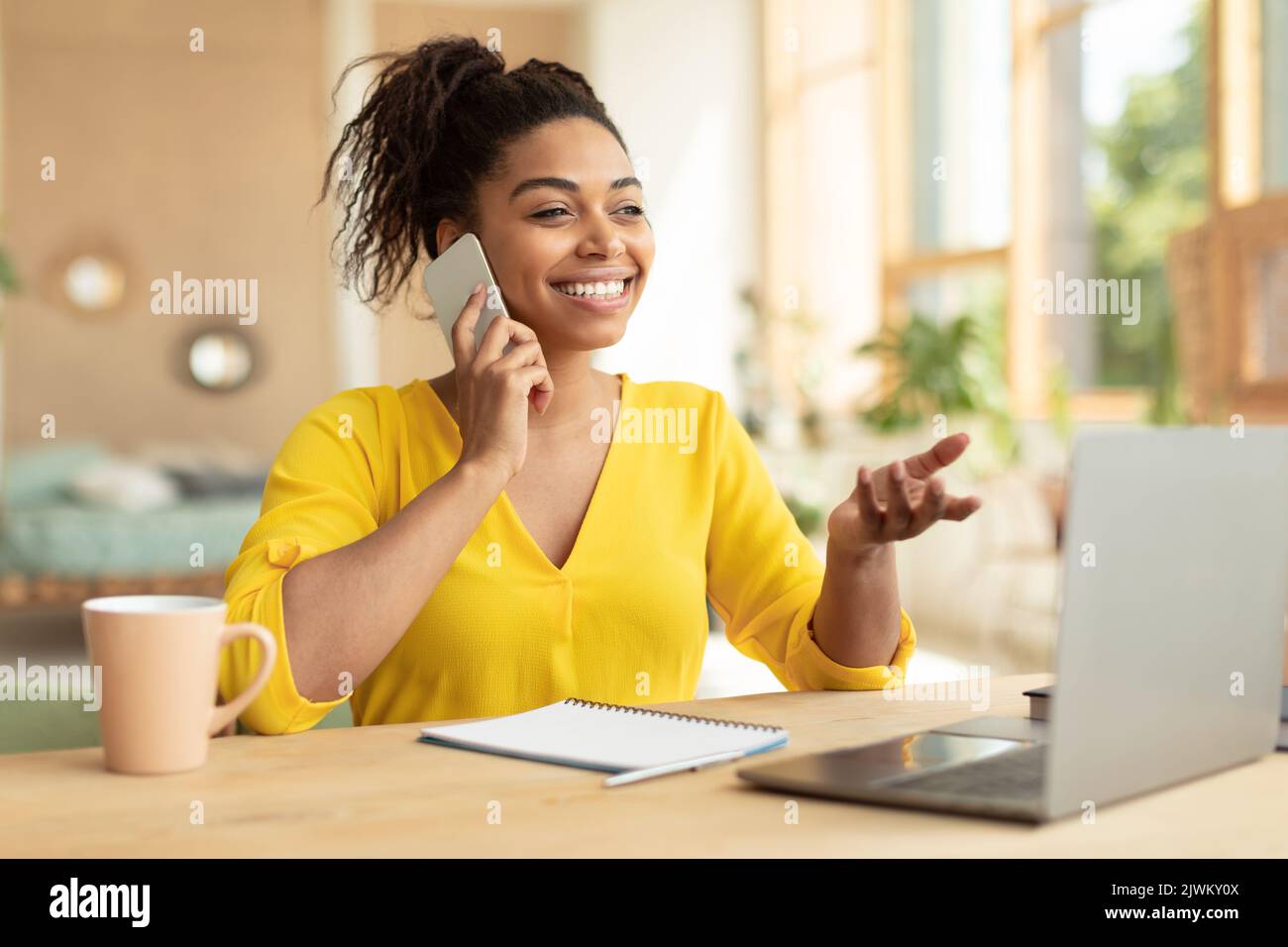 Portrait d'une femme noire excitée travaillant à la maison, parlant sur un smartphone et utilisant un ordinateur portable, assis au bureau à la maison Banque D'Images