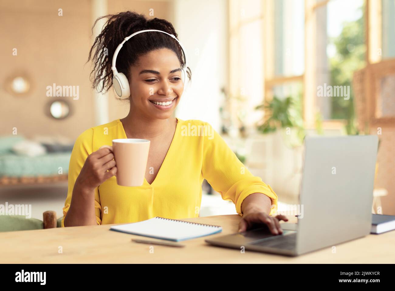Bonne femme afro-américaine qui boit du café et qui travaille sur un ordinateur portable, qui porte un casque, qui est assise au bureau Banque D'Images