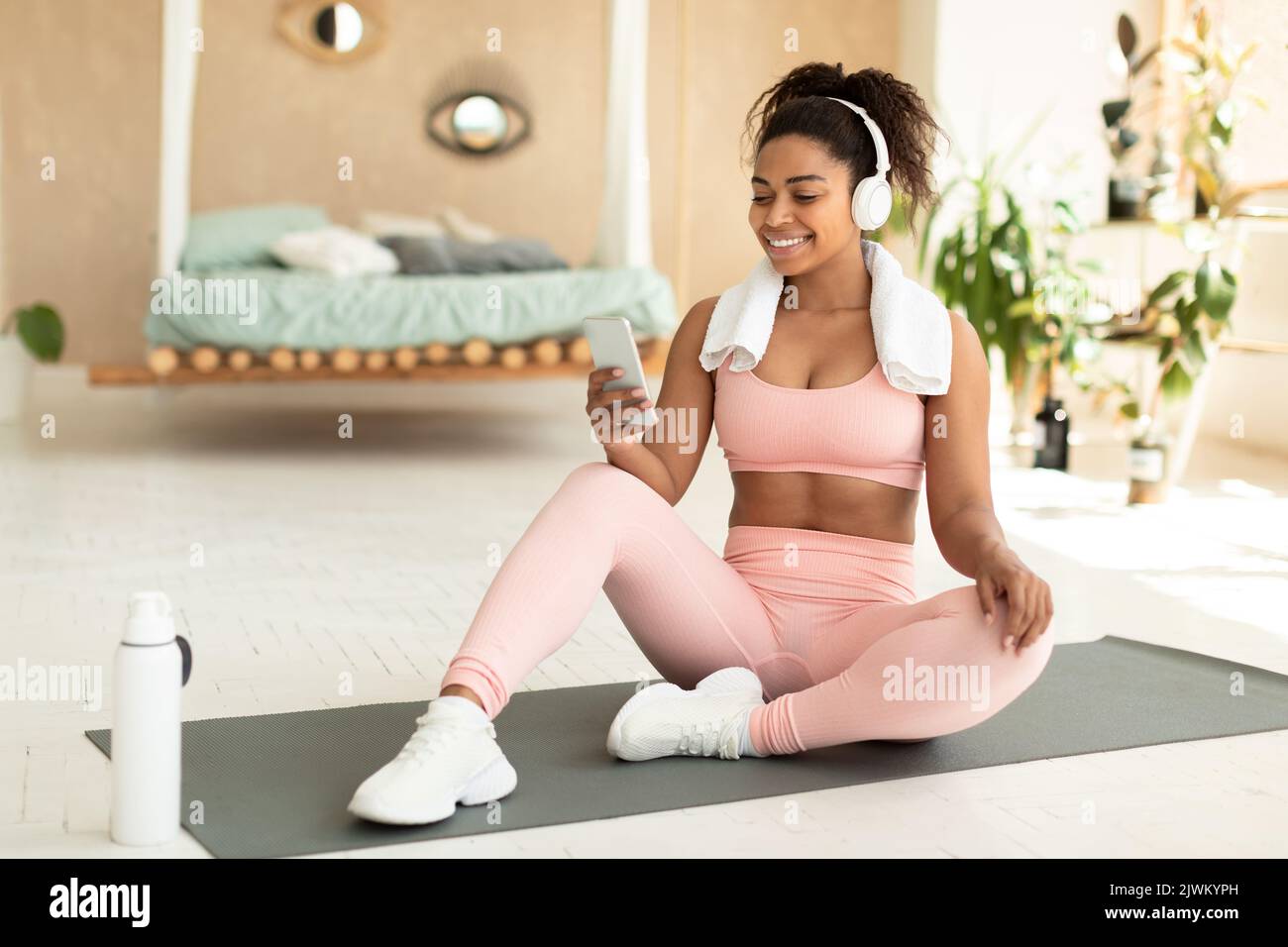 Femme noire à la coupe positive avec une serviette sur les épaules et un casque sans fil assis sur un tapis de fitness et écoutant de la musique Banque D'Images