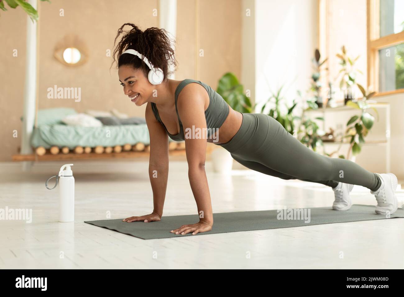 Restez au fitness à la maison. Bonne dame noire faisant l'entraînement de corps entier, debout dans la posture de planche et d'écouter de la musique dans le casque Banque D'Images