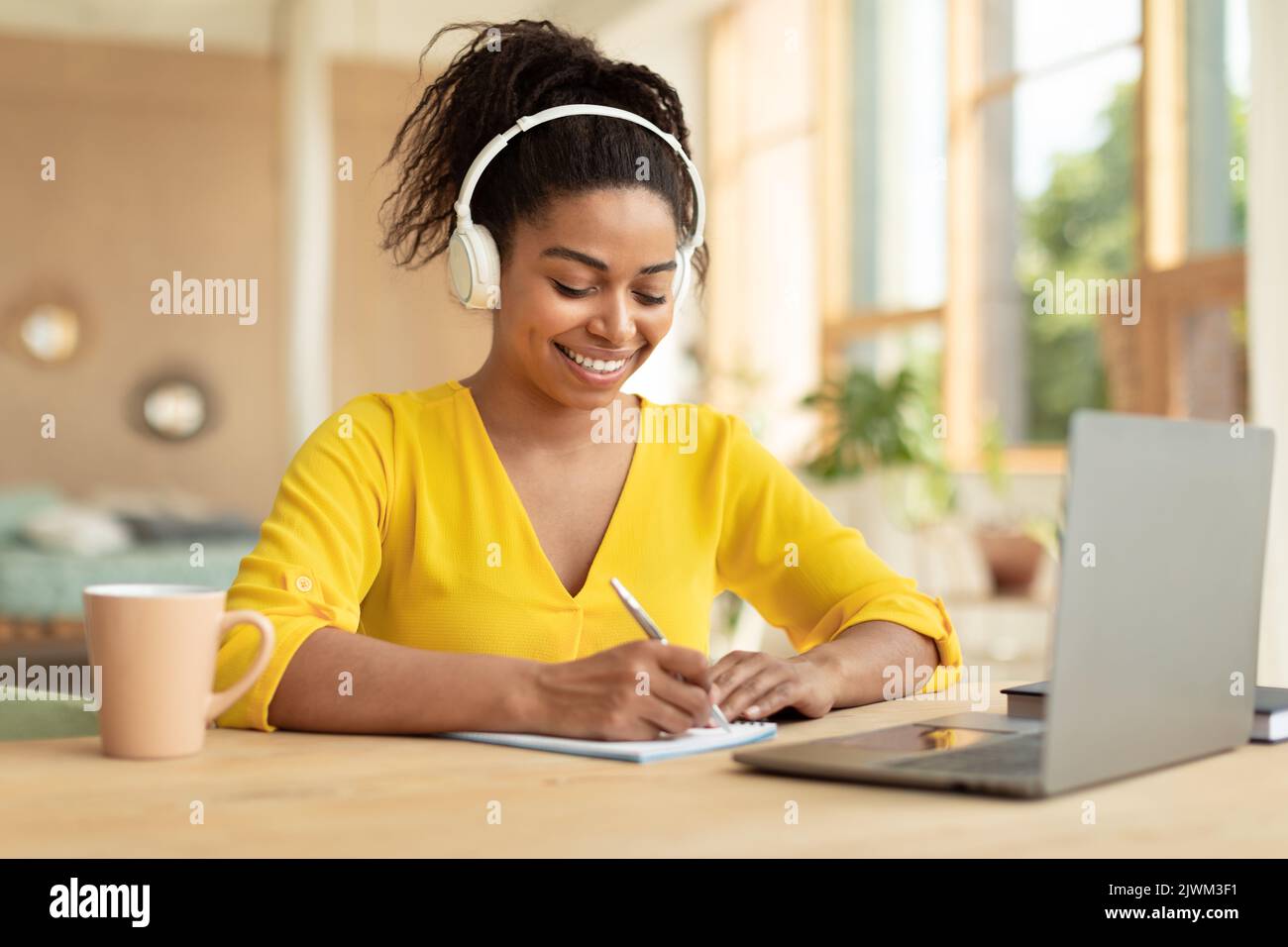 Bonne dame afro-américaine qui étudie à distance depuis la maison, utilise un ordinateur portable et prend des notes, assise au bureau à la maison Banque D'Images