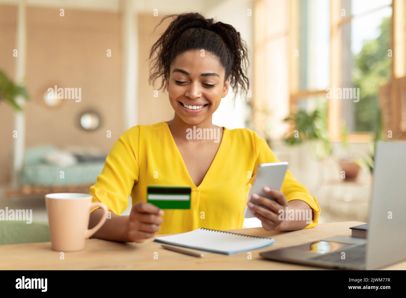 Femme américaine africaine positive avec smartphone et carte de crédit achetant des biens sur le Web, assis au bureau à la maison Banque D'Images